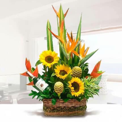 Arreglo floral tropical con girasoles amarillos, aves del paraíso naranjas, follaje verde y una botella de vino blanco en una canasta de mimbre sobre fondo interior blanco