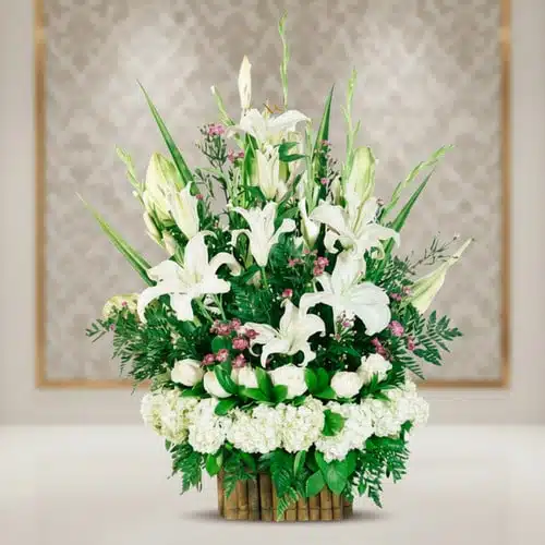 Arreglo floral con lirios blancos y follaje verde Arreglo floral elegante con lirios blancos, rosas blancas y follaje verde en base de bambú sobre fondo desenfocado