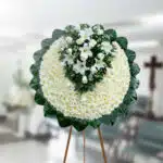 Corona fúnebre redonda de flores blancas con centro de lirios y rosas blancas, montada sobre caballete de madera en una sala velatoria