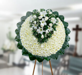 Corona fúnebre redonda de flores blancas con centro de lirios y rosas blancas, montada sobre caballete de madera en una sala velatoria
