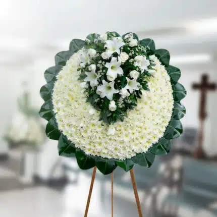 Corona fúnebre redonda de flores blancas con centro de lirios y rosas blancas, montada sobre caballete de madera en una sala velatoria