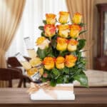 Arreglo floral de rosas amarillas y naranjas con dos botellas de cerveza clara en base rectangular blanca con lazo beige sobre mesa de madera