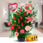 Arreglo floral en maceta negra con rosas rojas, lirios rosados, gerberas y globo rojo en forma de corazón con texto Te amo junto a caja de chocolates Ferrero Rocher