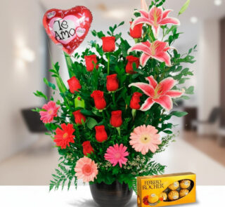 Arreglo floral en maceta negra con rosas rojas, lirios rosados, gerberas y globo rojo en forma de corazón con texto Te amo junto a caja de chocolates Ferrero Rocher
