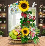 Arreglo de cumpleaños en canasta de mimbre con girasoles, rosas rojas, lirios rosados, frutas variadas y globo con mensaje Feliz Cumpleaños sobre mesa de madera