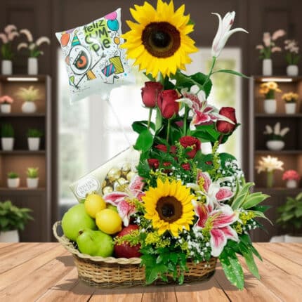 Arreglo de cumpleaños en canasta de mimbre con girasoles, rosas rojas, lirios rosados, frutas variadas y globo con mensaje Feliz Cumpleaños sobre mesa de madera