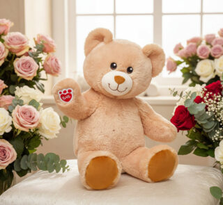 Osito de peluche beige sentado sobre un cojín blanco, rodeado de ramos de rosas rosas, rojas y blancas frente a una ventana