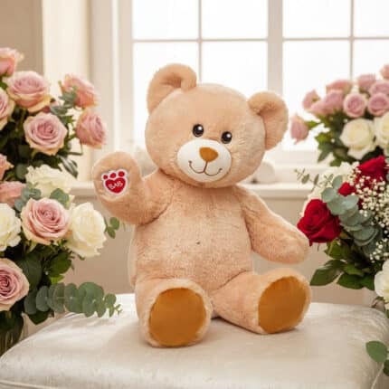 Osito de peluche beige sentado sobre un cojín blanco, rodeado de ramos de rosas rosas, rojas y blancas frente a una ventana