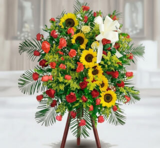 Arreglo funerario de pie con girasoles amarillos, rosas rojas y follaje verde sobre caballete de madera