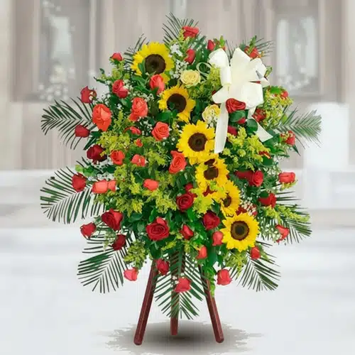 Arreglo funerario de pie con girasoles amarillos, rosas rojas y follaje verde sobre caballete de madera