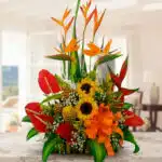 Arreglo floral tropical sobre mesa de mármol con girasoles, anturios rojos, heliconias y lazos naranjas en interior iluminado