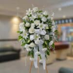 Arreglo floral grande de flores blancas con lazos y follaje verde sobre caballete de madera en una sala interior desenfocada