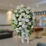 Arreglo floral grande de flores blancas con lazos y follaje verde sobre caballete de madera en una sala interior desenfocada