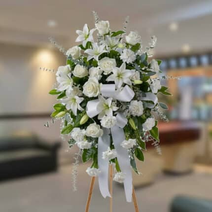 Arreglo floral grande de flores blancas con lazos y follaje verde sobre caballete de madera en una sala interior desenfocada