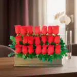 Arreglo de varias rosas rojas en filas dentro de una caja de madera con hojas verdes, colocado sobre una mesa en interior