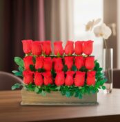 Arreglo de varias rosas rojas en filas dentro de una caja de madera con hojas verdes, colocado sobre una mesa en interior