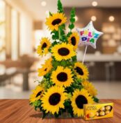 Arreglo floral de girasoles amarillos en forma piramidal sobre mesa de madera, con globo de estrella que dice feliz día y caja de chocolates Ferrero Rocher al frente, en interior desenfocado