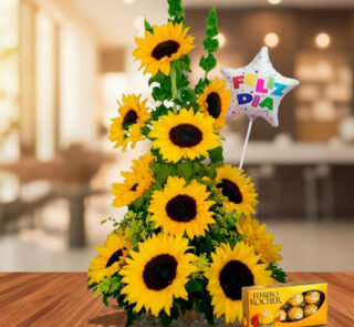 Arreglo floral de girasoles amarillos en forma piramidal sobre mesa de madera, con globo de estrella que dice feliz día y caja de chocolates Ferrero Rocher al frente, en interior desenfocado