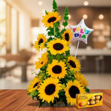 Arreglo floral de girasoles amarillos en forma piramidal sobre mesa de madera, con globo de estrella que dice feliz día y caja de chocolates Ferrero Rocher al frente, en interior desenfocado