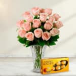 Arreglo de rosas rosadas en un florero de cristal transparente acompañado de una caja de chocolates Ferrero Rocher sobre superficie de mármol