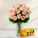 Arreglo de rosas rosadas en un florero de cristal transparente acompañado de una caja de chocolates Ferrero Rocher sobre superficie de mármol