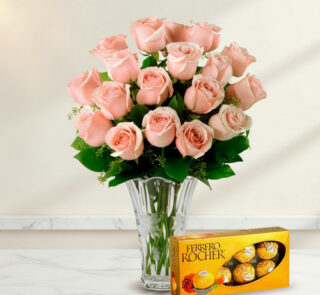 Arreglo de rosas rosadas en un florero de cristal transparente acompañado de una caja de chocolates Ferrero Rocher sobre superficie de mármol