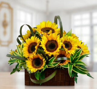 Arreglo floral de girasoles amarillos con follaje verde en una cesta cuadrada de madera sobre una mesa, con fondo de interior desenfocado