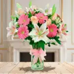 Arreglo floral con lirios blancos y rosas, gerberas rosas y rosas melocotón en jarrón de cristal con lazo rosa sobre mesa de madera