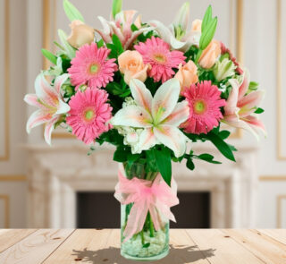 Arreglo floral con lirios blancos y rosas, gerberas rosas y rosas melocotón en jarrón de cristal con lazo rosa sobre mesa de madera