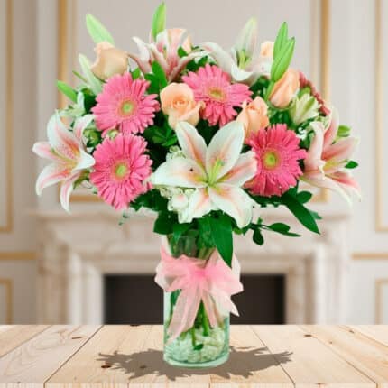 Arreglo floral con lirios blancos y rosas, gerberas rosas y rosas melocotón en jarrón de cristal con lazo rosa sobre mesa de madera