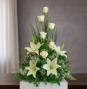 Arreglo floral vertical con rosas blancas, lirios blancos y follaje verde sobre una mesa de madera frente a fondo gris