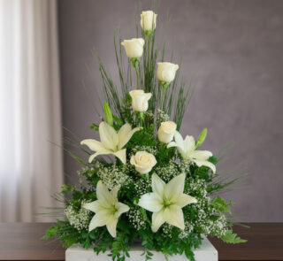 Arreglo floral vertical con rosas blancas, lirios blancos y follaje verde sobre una mesa de madera frente a fondo gris