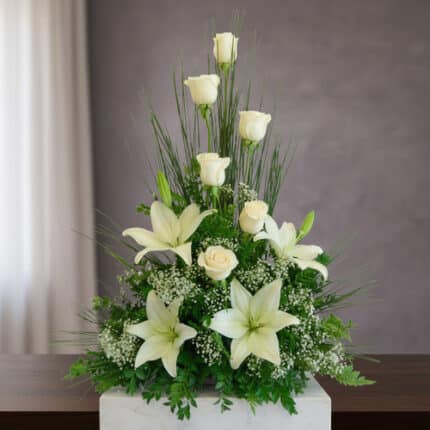 Arreglo floral vertical con rosas blancas, lirios blancos y follaje verde sobre una mesa de madera frente a fondo gris
