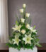 Arreglo floral vertical con rosas blancas, lirios blancos y follaje verde sobre una mesa de madera frente a fondo gris