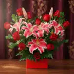 Arreglo de lirios orientales rosados y rosas rojas en caja roja sobre mesa de madera con cortina burdeos de fondo