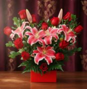Arreglo de lirios orientales rosados y rosas rojas en caja roja sobre mesa de madera con cortina burdeos de fondo