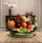 Canasta tejida con variedad de frutas frescas como manzanas, uvas, mangos y cítricos sobre una mesa de madera, con fondo de sala elegante desenfocado