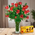Arreglo de rosas rojas en jarrón de cristal junto a una caja de chocolates Ferrero Rocher sobre una mesa de madera en sala iluminada