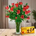 Arreglo de rosas rojas en jarrón de cristal junto a una caja de chocolates Ferrero Rocher sobre una mesa de madera en sala iluminada