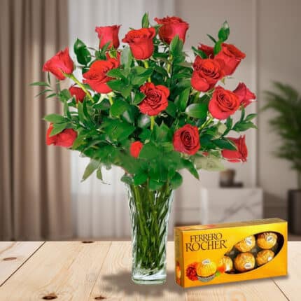 Arreglo de rosas rojas en jarrón de cristal junto a una caja de chocolates Ferrero Rocher sobre una mesa de madera en sala iluminada