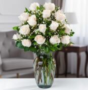 Arreglo de rosas blancas con follaje verde y paniculata en un jarrón de cristal sobre una mesa en una sala elegante