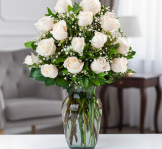 Arreglo de rosas blancas con follaje verde y paniculata en un jarrón de cristal sobre una mesa en una sala elegante