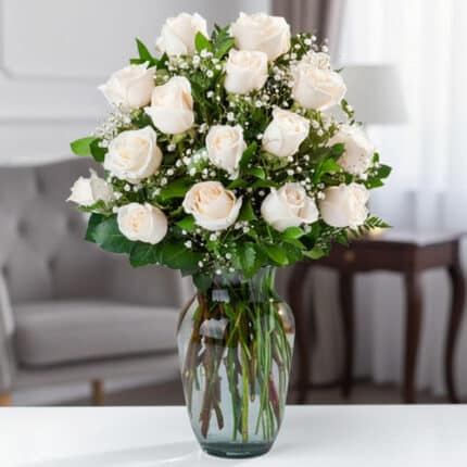 Arreglo de rosas blancas con follaje verde y paniculata en un jarrón de cristal sobre una mesa en una sala elegante