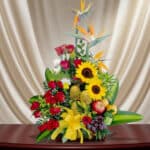 Arreglo floral colorido con girasoles amarillos, rosas rojas, aves del paraíso y frutas como uvas y manzanas sobre una mesa de madera, con fondo de cortina beige