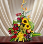 Arreglo floral colorido con girasoles amarillos, rosas rojas, aves del paraíso y frutas como uvas y manzanas sobre una mesa de madera, con fondo de cortina beige