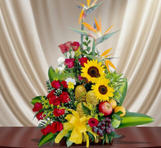 Arreglo floral colorido con girasoles amarillos, rosas rojas, aves del paraíso y frutas como uvas y manzanas sobre una mesa de madera, con fondo de cortina beige
