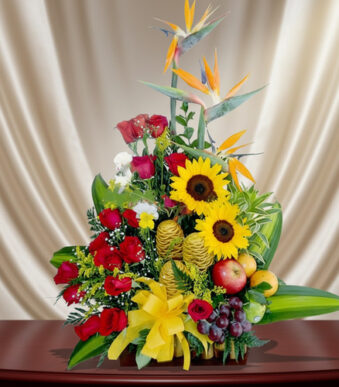 Arreglo floral colorido con girasoles amarillos, rosas rojas, aves del paraíso y frutas como uvas y manzanas sobre una mesa de madera, con fondo de cortina beige