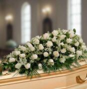 Centro fúnebre alargado de rosas blancas y follaje verde colocado sobre un ataúd de madera clara en una sala iluminada