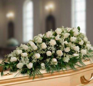 Centro fúnebre alargado de rosas blancas y follaje verde colocado sobre un ataúd de madera clara en una sala iluminada