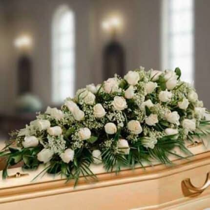 Centro fúnebre alargado de rosas blancas y follaje verde colocado sobre un ataúd de madera clara en una sala iluminada